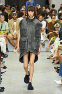 Model walking a runway in a oversized gray denim vest-dress with large front pockets and a high-low hem runway look.