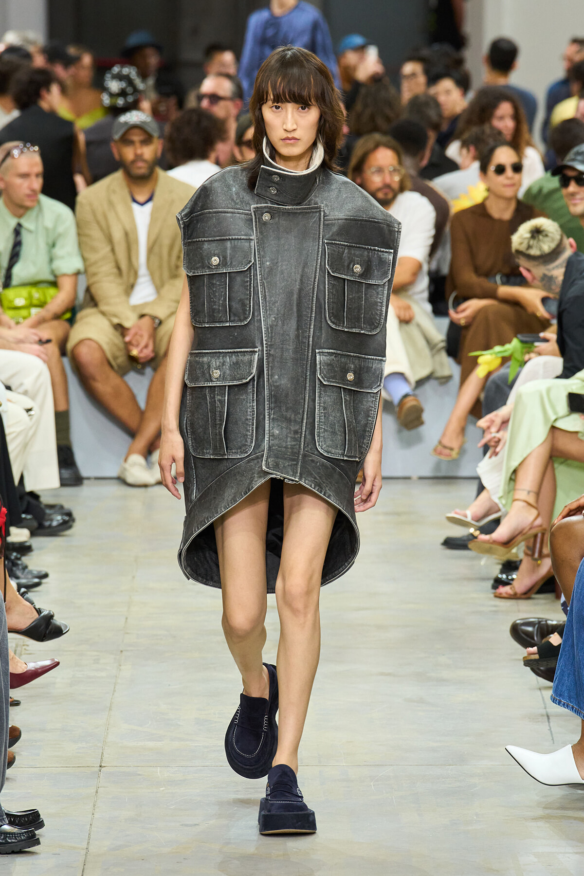 Model walking a runway in a oversized gray denim vest-dress with large front pockets and a high-low hem runway look.