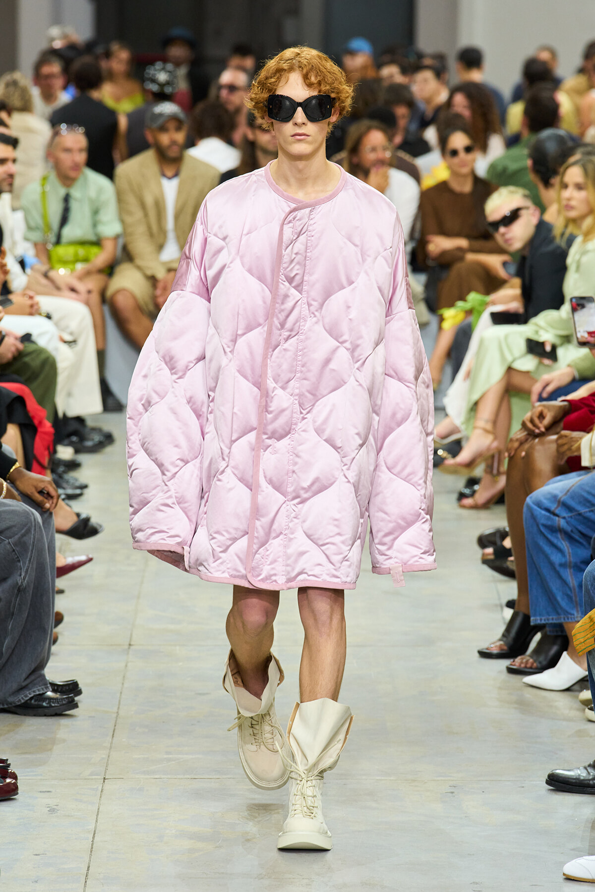 Model on a runway wearing an oversized quilted pink coat, large black sunglasses, and cream boots as an audience watches.