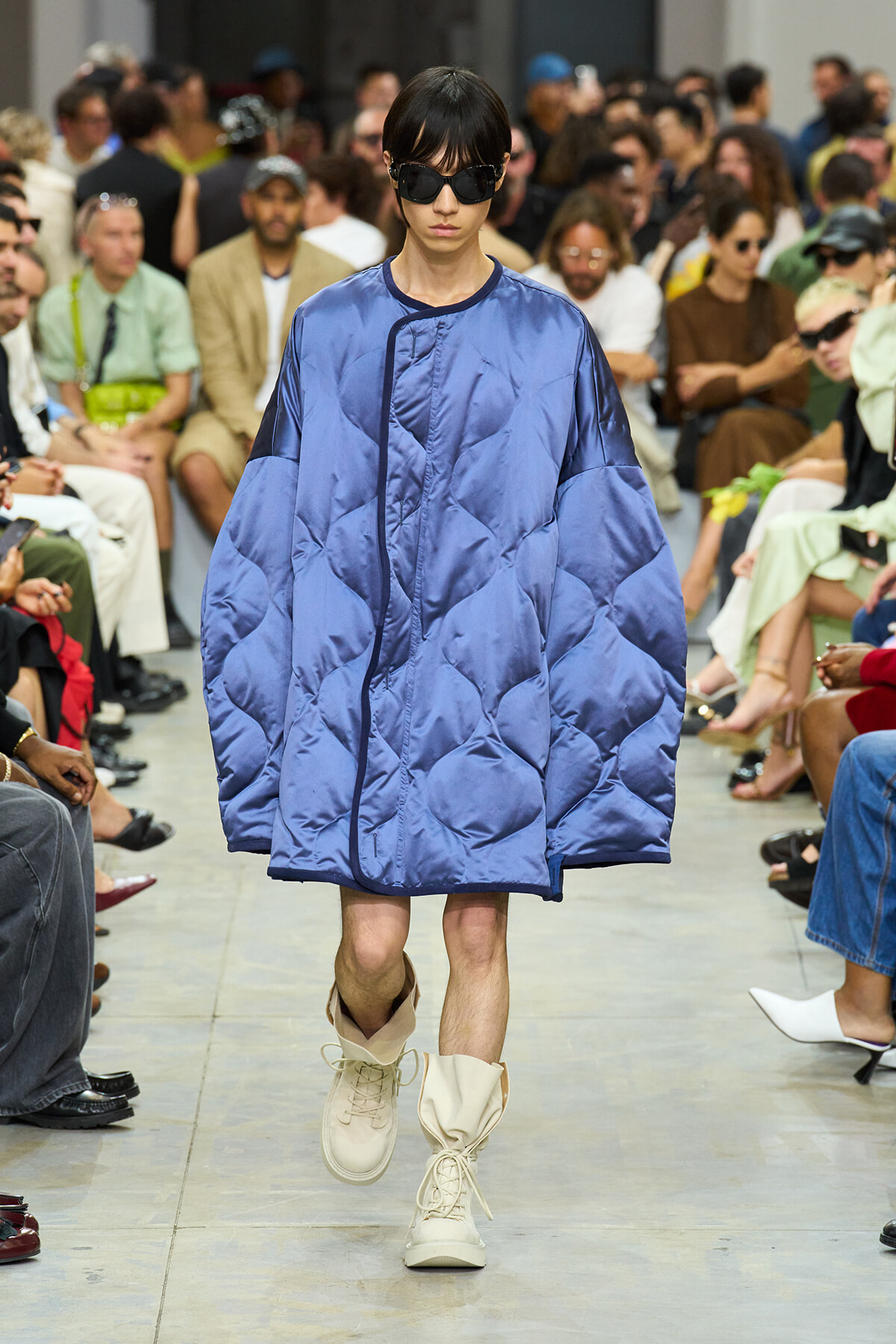 Model walks the runway wearing an oversized blue quilted coat, black sunglasses, and cream boots on a crowded fashion show runway.
