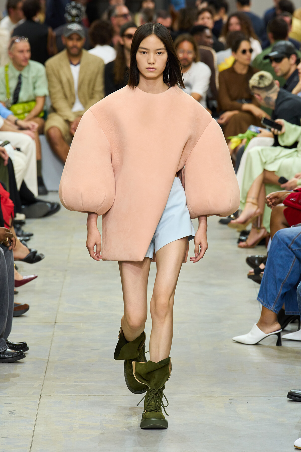 Model walks the runway in an oversized blush pink coat with balloon sleeves over a pale blue dress, paired with olive green combat boots.