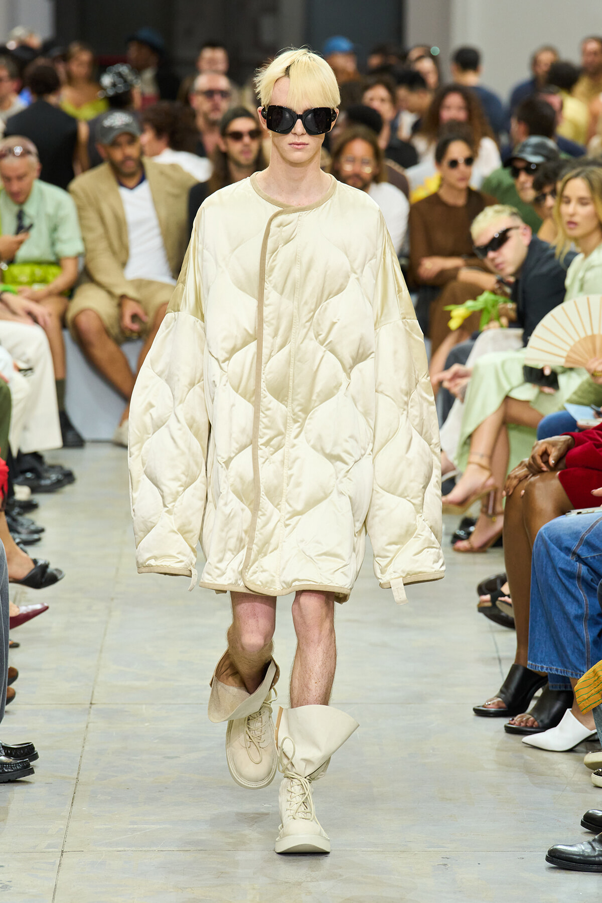 Model walking the runway in an oversized cream quilted coat with matching boots and large black sunglasses, audience in the background.