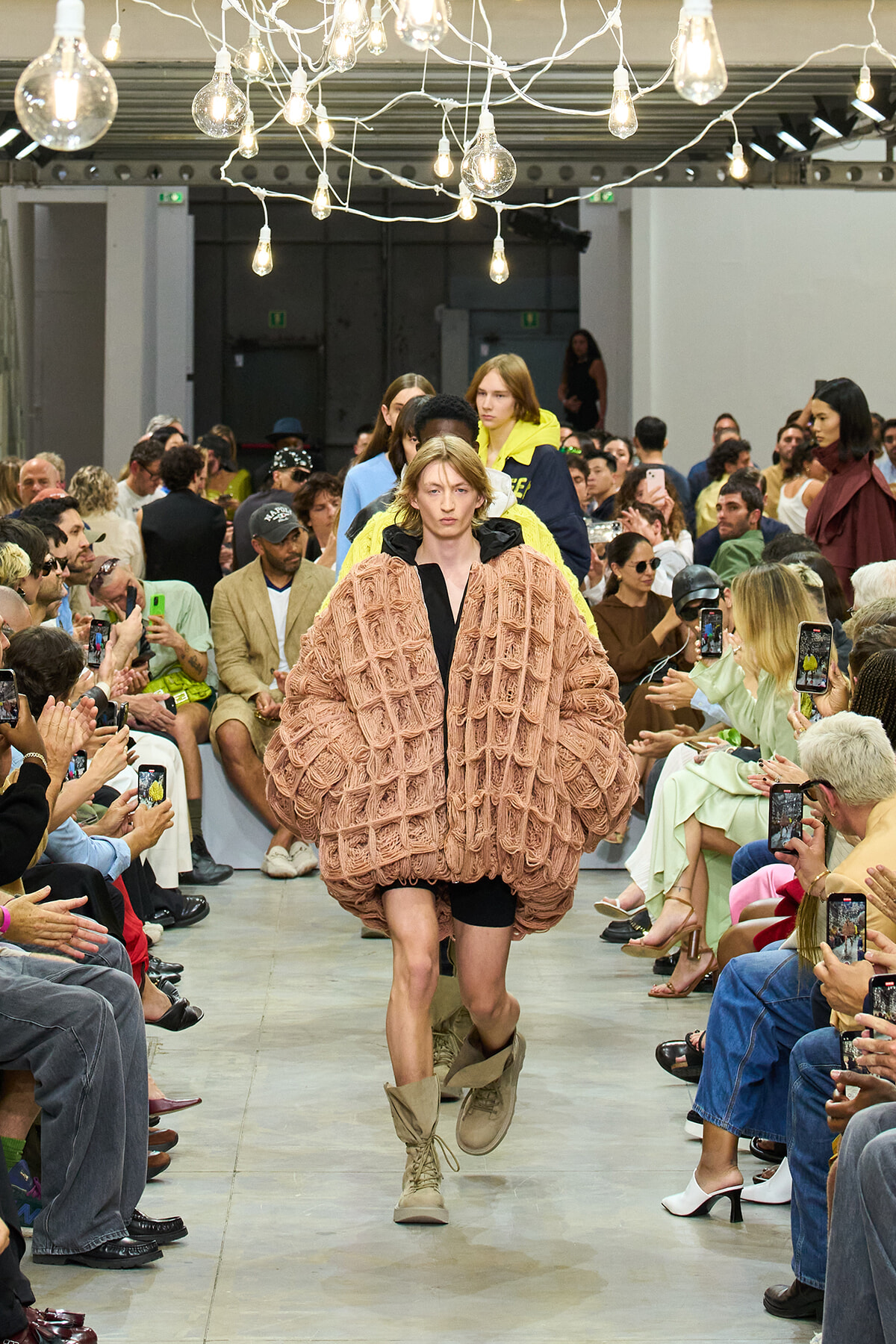 Model walks a runway in an oversized pink quilted coat with beige boots; audience on both sides watches and films the show.