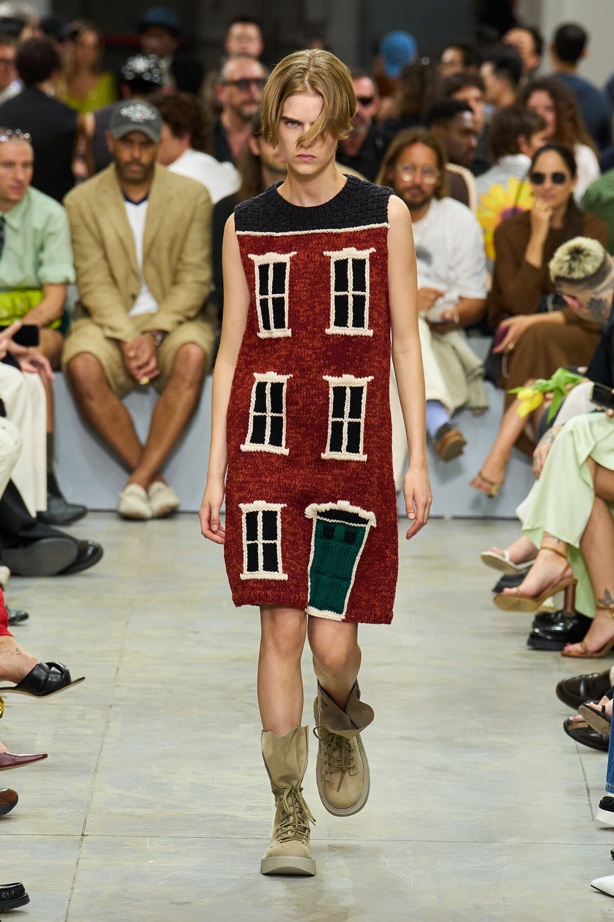 Model walking the runway in a sleeveless red dress knitted to look like a house facade with white-framed windows and a green door, paired with tan combat boots.