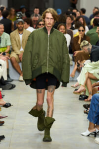 Model walks the runway in an oversized olive bomber jacket, black shorts, and green combat boots as the audience watches.