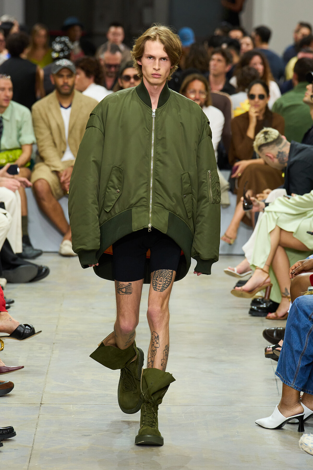 Model walks the runway in an oversized olive bomber jacket, black shorts, and green combat boots as the audience watches.