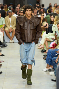 Male model walks the runway in an oversized brown leather jacket with broad shoulders, paired with loose light-wash denim shorts and green combat boots, audience in background.