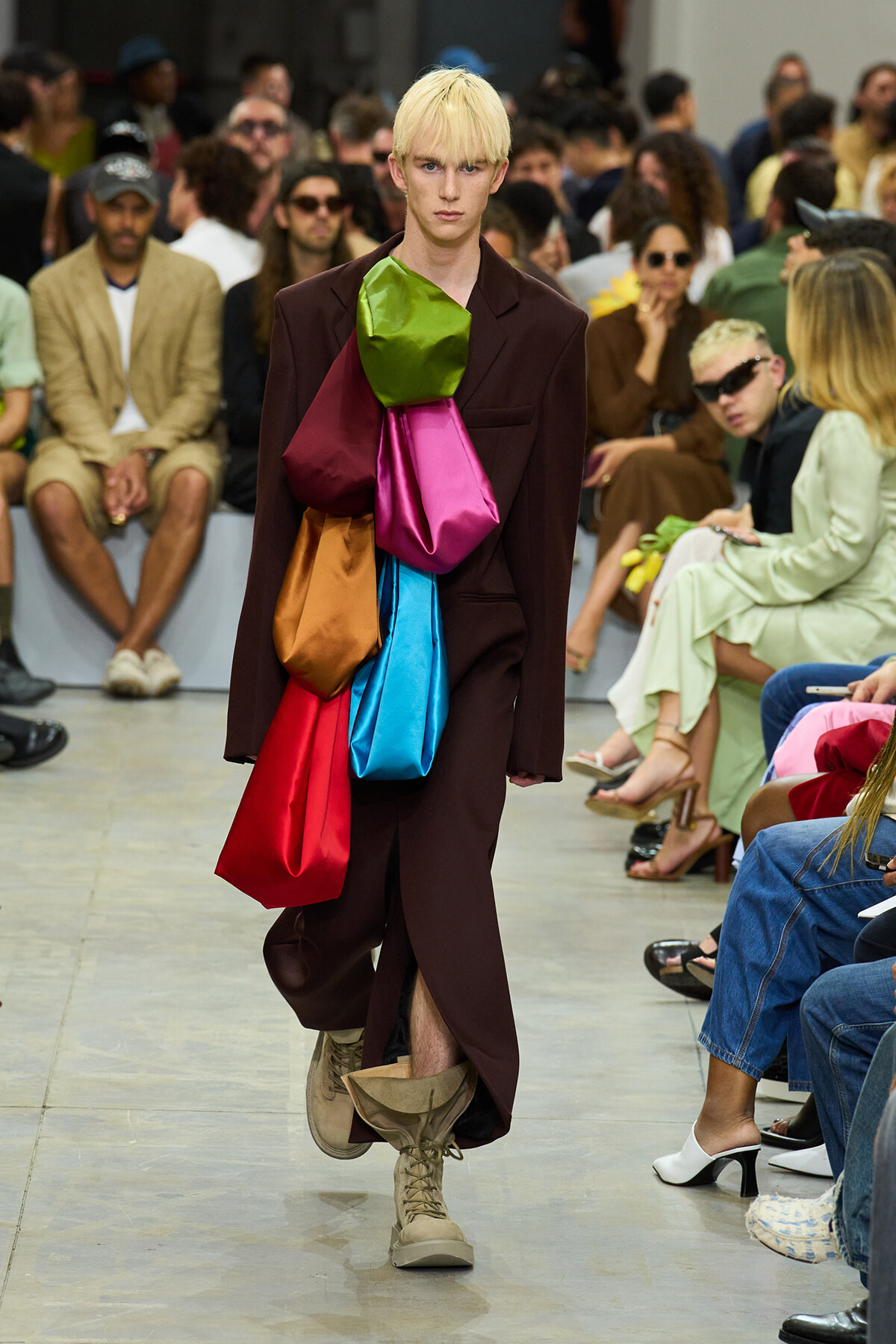 Model on a runway wearing a burgundy suit with oversized colorful fabric pouches (green, pink, blue, red, orange) attached to the chest and waist.