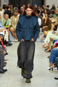 Model walks the runway in a blue oversized jacket with bold shoulders, paired with loose gray denim and olive-green boots; audience watches in the background.