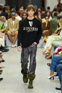 Male model walks a runway in a black Guinness sweater, gray pants, and olive-green boots; audience in the background.