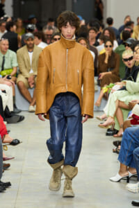 Model walks a fashion show in a tan oversized leather jacket, blue pants, and tan boots, with a seated audience in the background.