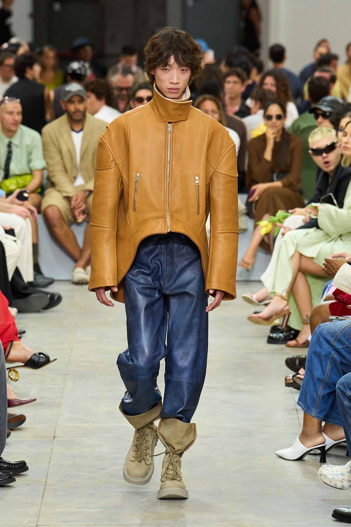 Model walks a fashion show in a tan oversized leather jacket, blue pants, and tan boots, with a seated audience in the background.