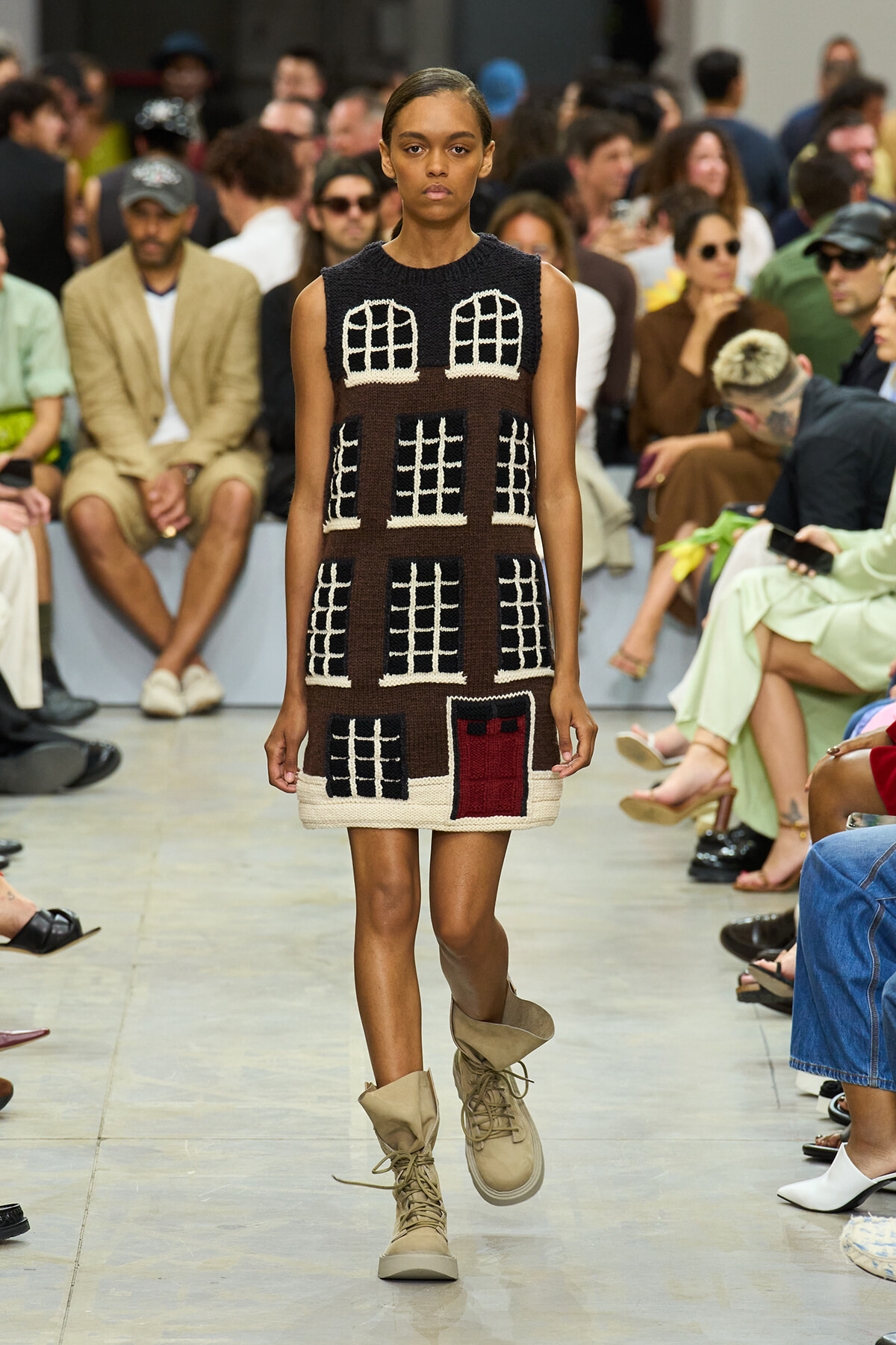 Model walks down runway wearing a sleeveless brown knit dress with white grid-window patterns and a red door pocket; beige combat boots add to the look.