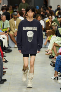 Model on a fashion runway wearing a navy oversized sweatshirt with 'GUINNESS' and a smiling pint graphic; beige boots, audience in the background.