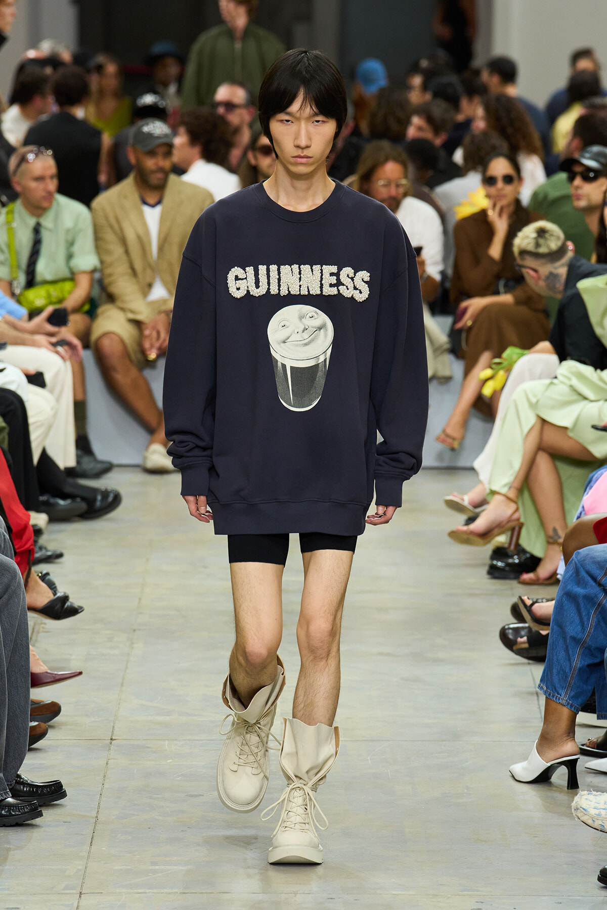 Model on a fashion runway wearing a navy oversized sweatshirt with 'GUINNESS' and a smiling pint graphic; beige boots, audience in the background.