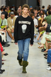 Male model walks a fashion show wearing a black Guinness sweatshirt, blue jeans, and green combat boots; audience sits along the runway.