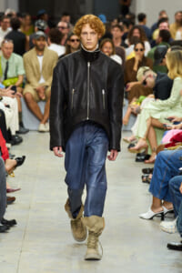 Model walks down runway in a black leather jacket, blue jeans, and tan combat boots with a crowd watching.