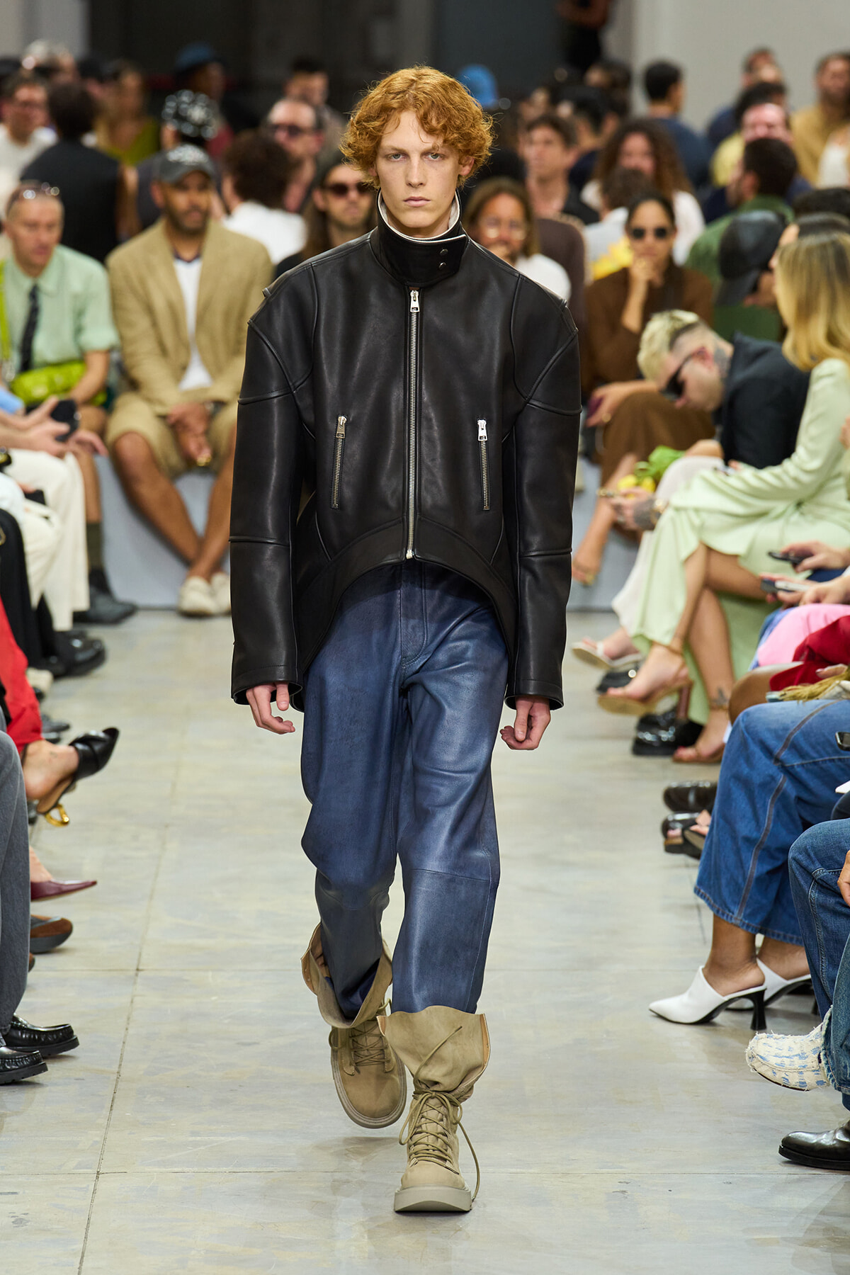 Model walks down runway in a black leather jacket, blue jeans, and tan combat boots with a crowd watching.