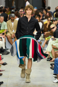 Male model walks a runway in a black button-down shirt with a large multicolored fabric wrap around the waist, beige boots, and a cheering audience in the background.