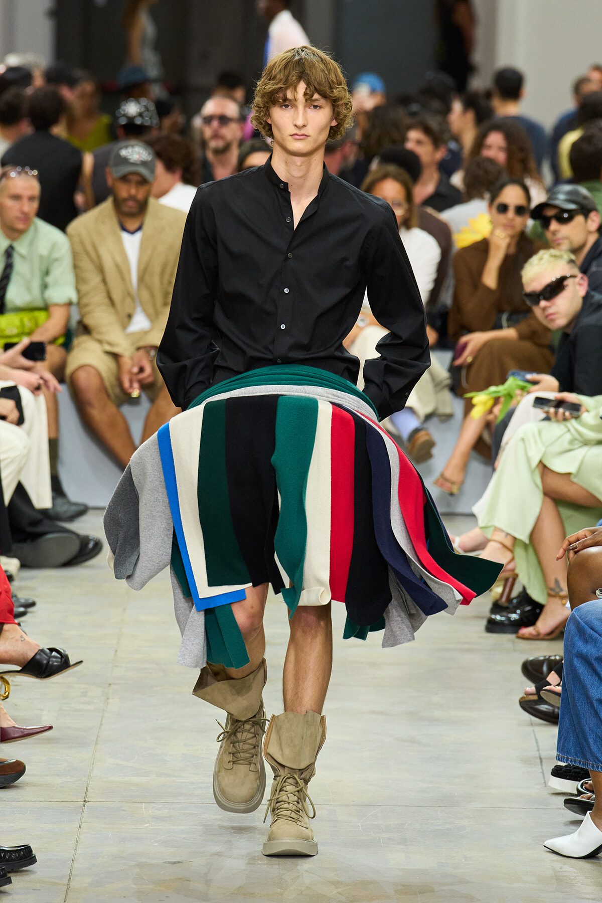 Male model walks a runway in a black button-down shirt with a large multicolored fabric wrap around the waist, beige boots, and a cheering audience in the background.