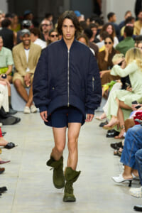 Model walks a fashion show in an oversized navy bomber jacket, navy shorts, and olive-green combat boots with a seated audience in the background.