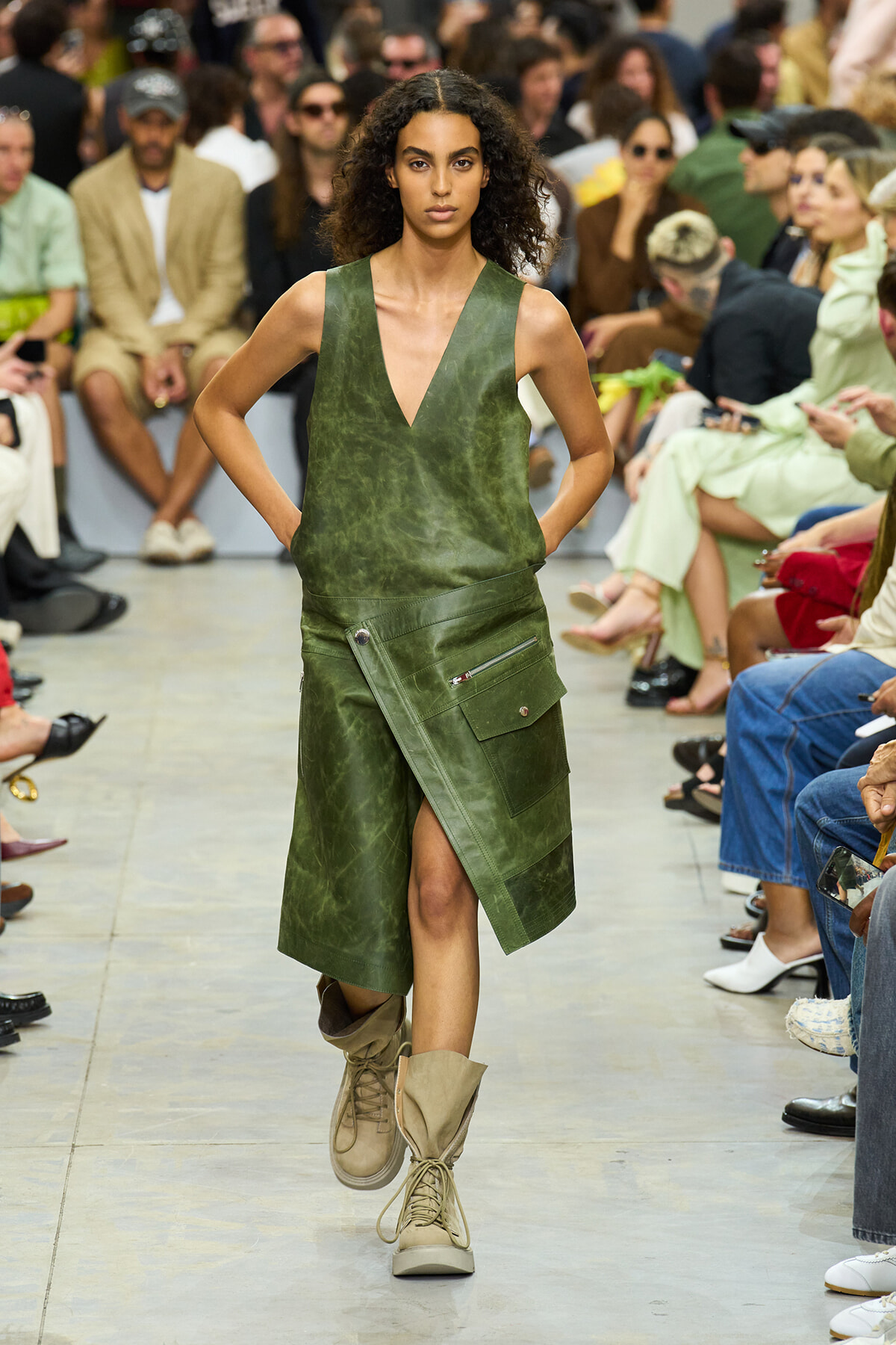 Model walks the runway in an olive-green sleeveless top and a matching asymmetrical cargo skirt with large pockets, wearing tan lace-up boots.