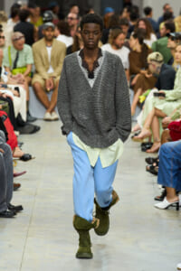 Model walks a fashion runway in a gray knit sweater with a lace V-neck, light yellow shirt underneath, light blue pants, and green combat boots.