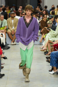 Model walks a fashion runway in a loose purple knit sweater over a black lace top and mint green pants with beige combat boots.