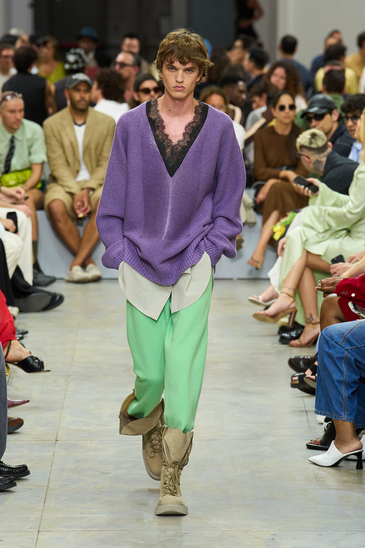 Model walks a fashion runway in a loose purple knit sweater over a black lace top and mint green pants with beige combat boots.