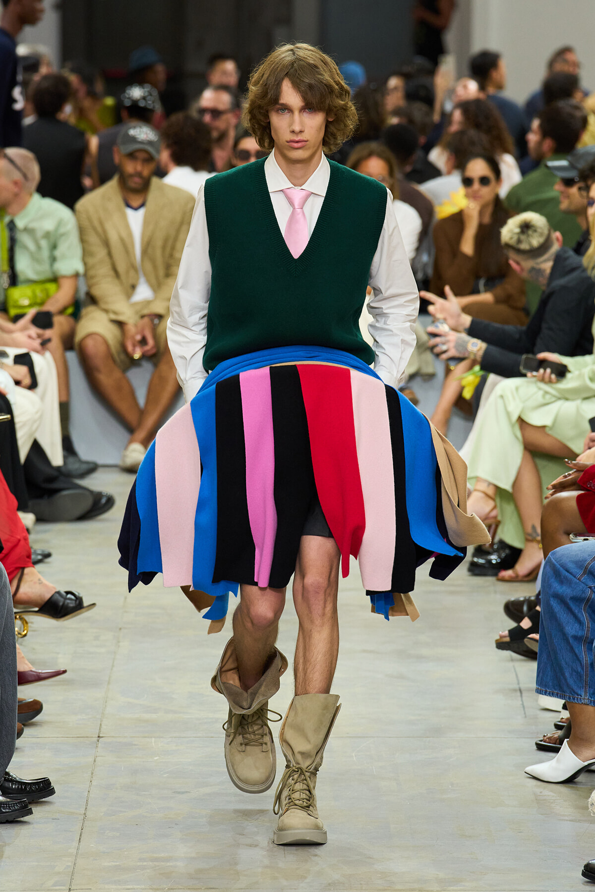 Male runway model walking the catwalk in a green V-neck sweater vest over a white shirt, pink tie, and a multi-color striped skirt; beige combat boots.