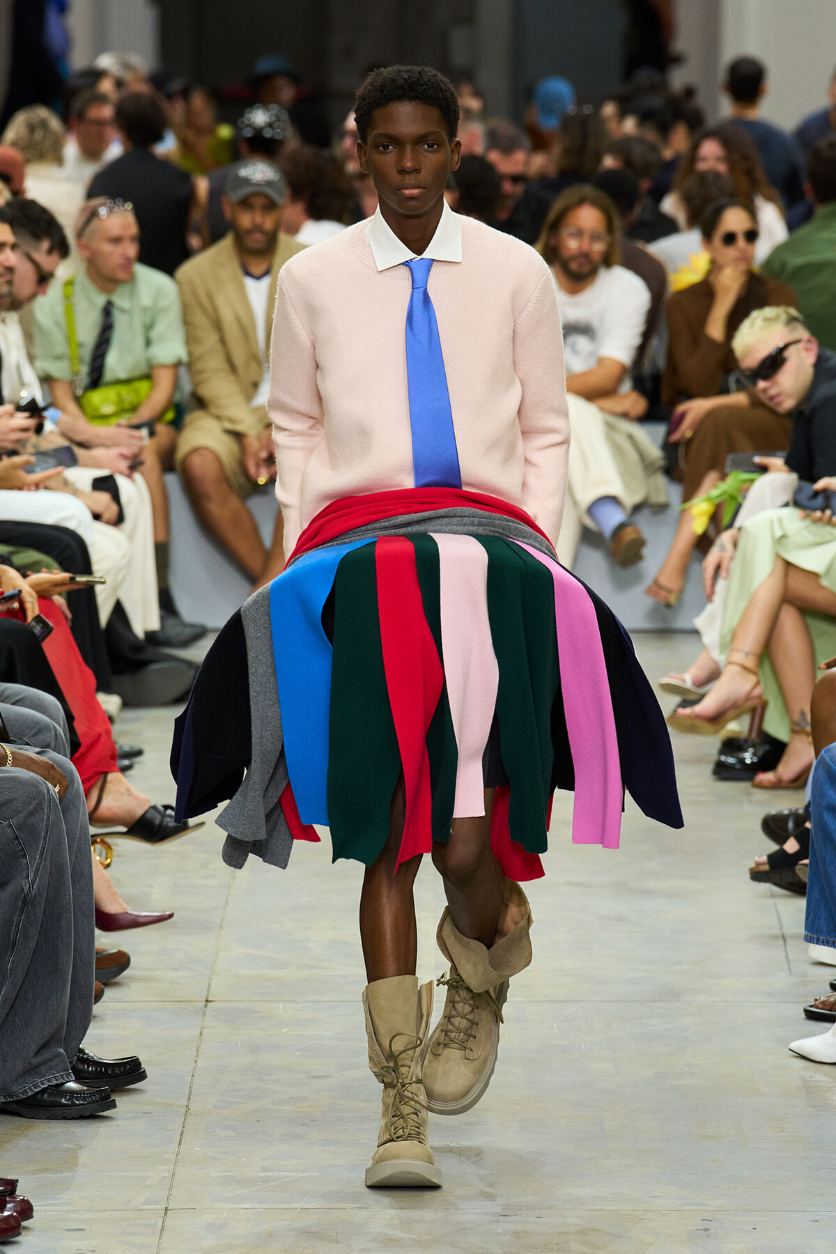 Male model walks a fashion show runway in a pale pink sweater with white collar and blue tie, plus a multicolored fringe skirt made of fabric strips; beige boots; audience in background