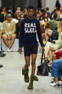 Model walking a runway wearing a navy sweater that reads 'REAL SLEE' paired with black shorts, green tall boots, and a black handbag; audience in background