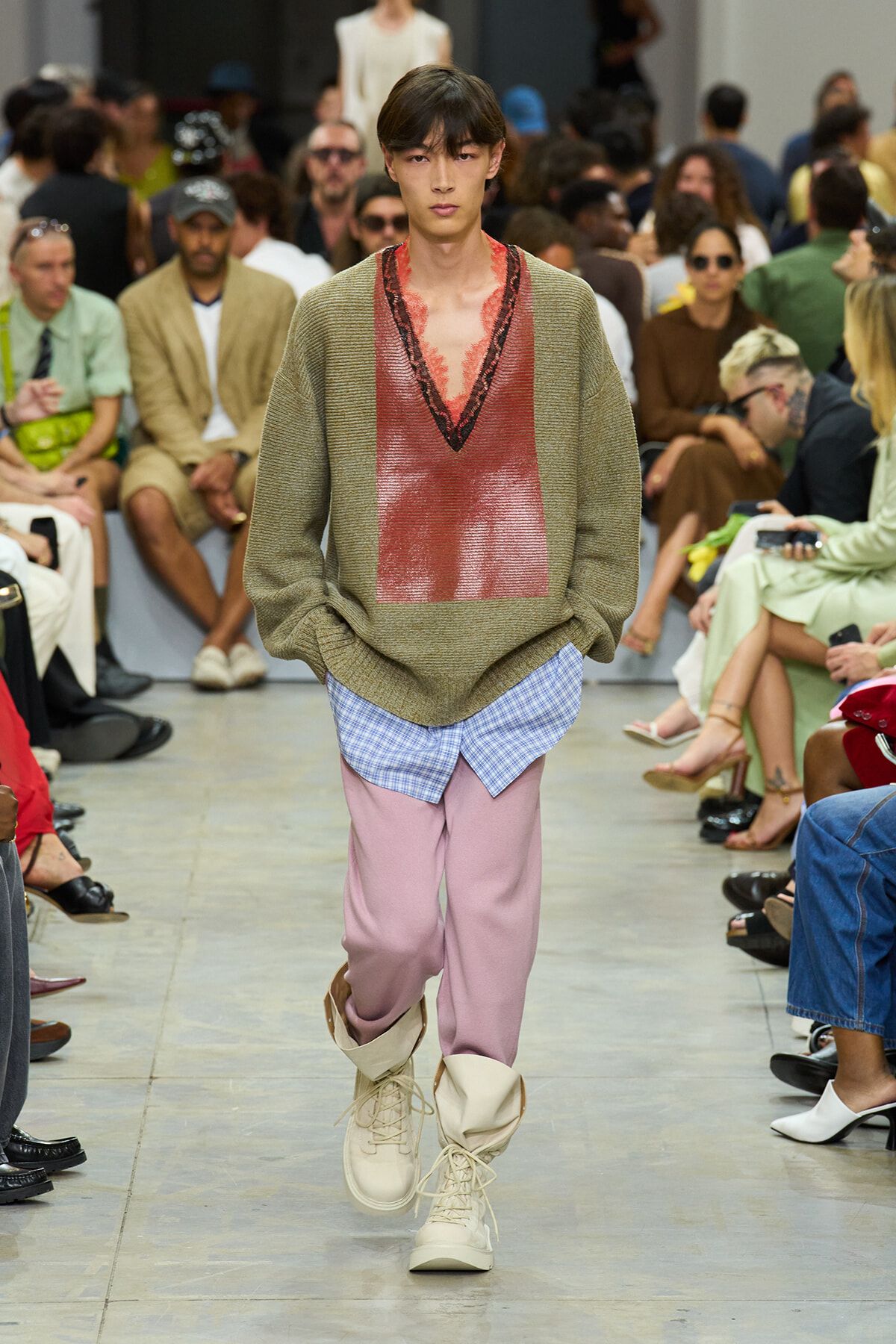Model on a runway wearing an oversized olive knit sweater with a red V-neck panel, pink pants, and beige lace-up boots.