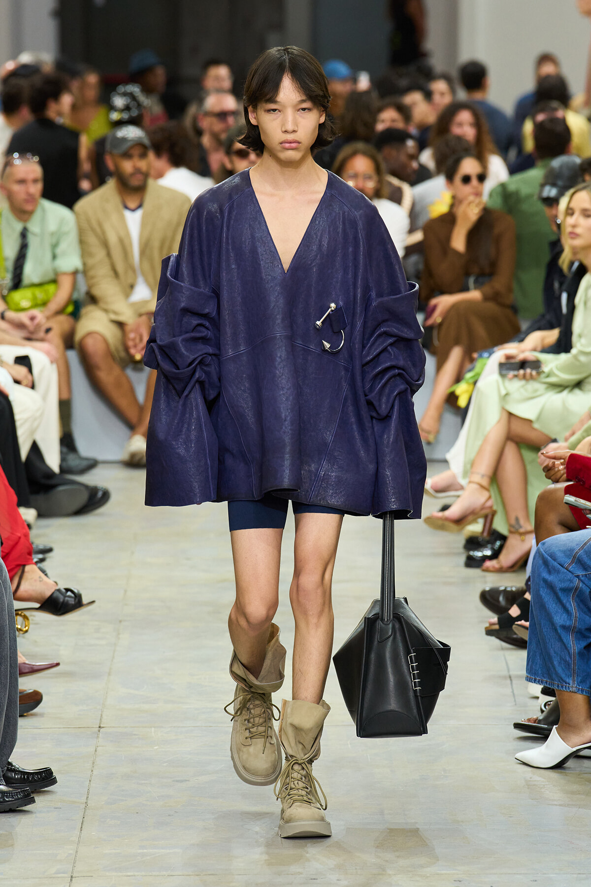 Model walking a runway in an oversized navy top with a deep V, short bottoms, tan combat boots, and a black geometric handbag.