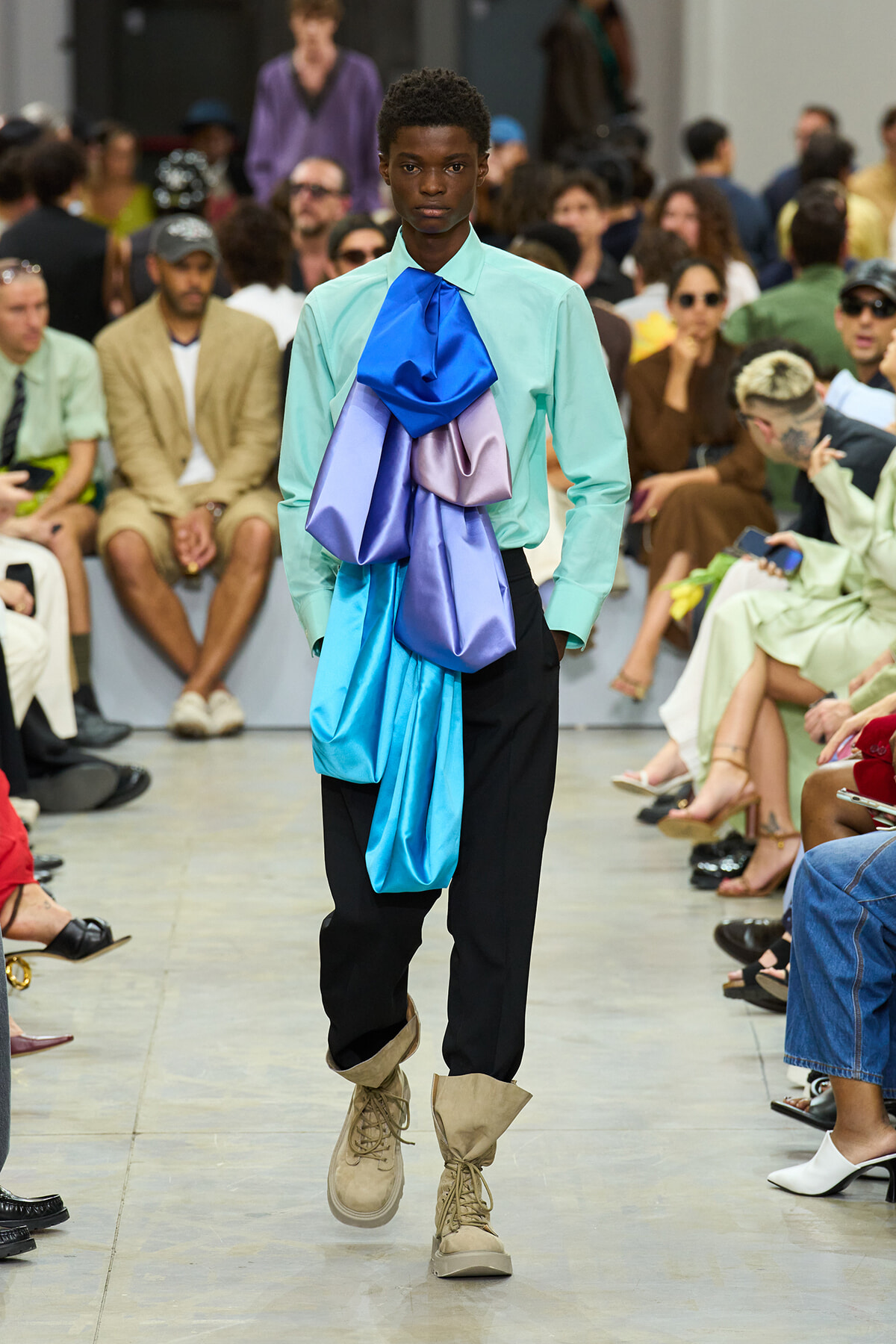 Male model on a runway wearing a mint shirt with a large blue and purple draped scarf and beige boots, with the audience in the background.