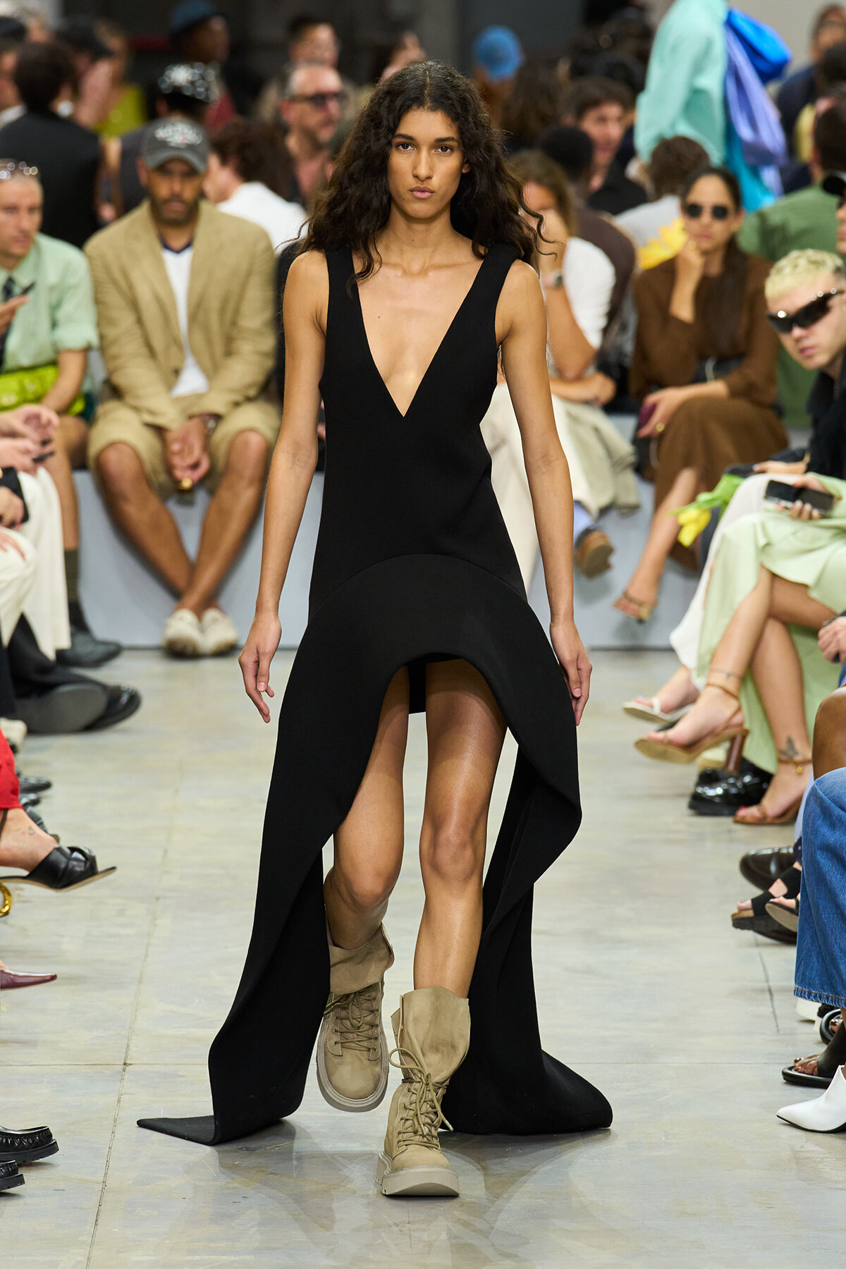 Runway model walks in a sleeveless black V-neck dress with a dramatic high-low hem, wearing beige combat boots as she strides forward.