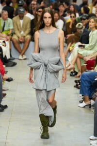 Model on a runway wearing a gray sleeveless dress with a large waist bow and olive-green lace-up boots.