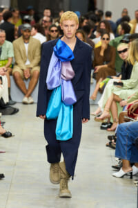 Model walks the runway wearing a navy suit with large blue and lavender fabric drapes cascading from the chest; tan boots and a focused audience in the background.