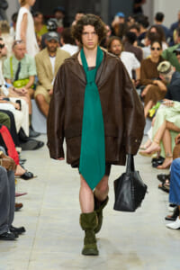 Model walks a fashion runway in an oversized brown leather coat, a bright green scarf tied as a necktie, green boots, and carries a black structured tote. Audience blurred in the background.