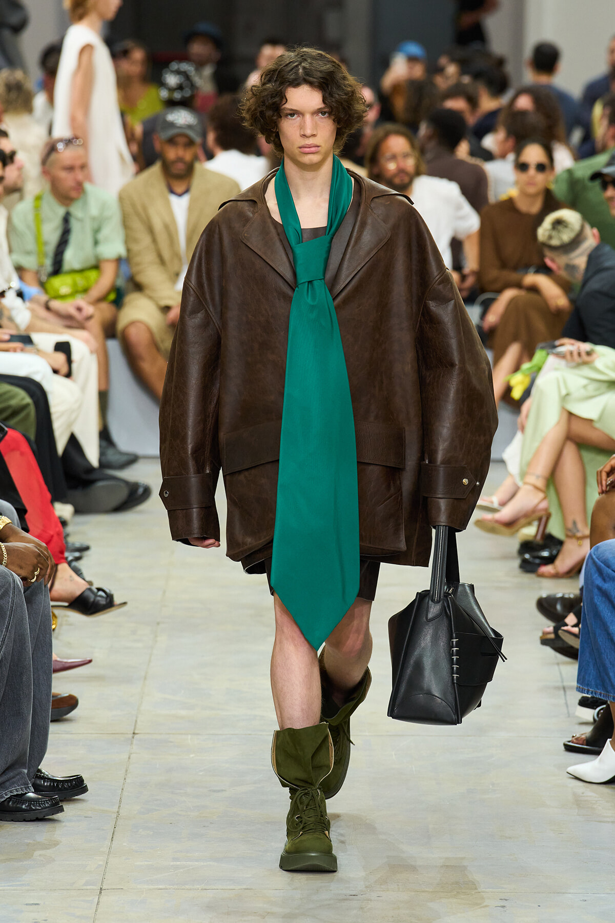 Model walks a fashion runway in an oversized brown leather coat, a bright green scarf tied as a necktie, green boots, and carries a black structured tote. Audience blurred in the background.