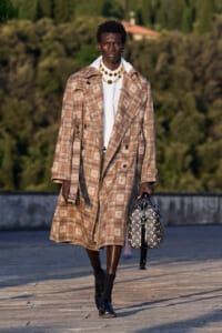 Model wearing a beige plaid trench coat over a white shirt, accessorized with gold necklaces and carrying a patterned handbag on a runway outdoor setting