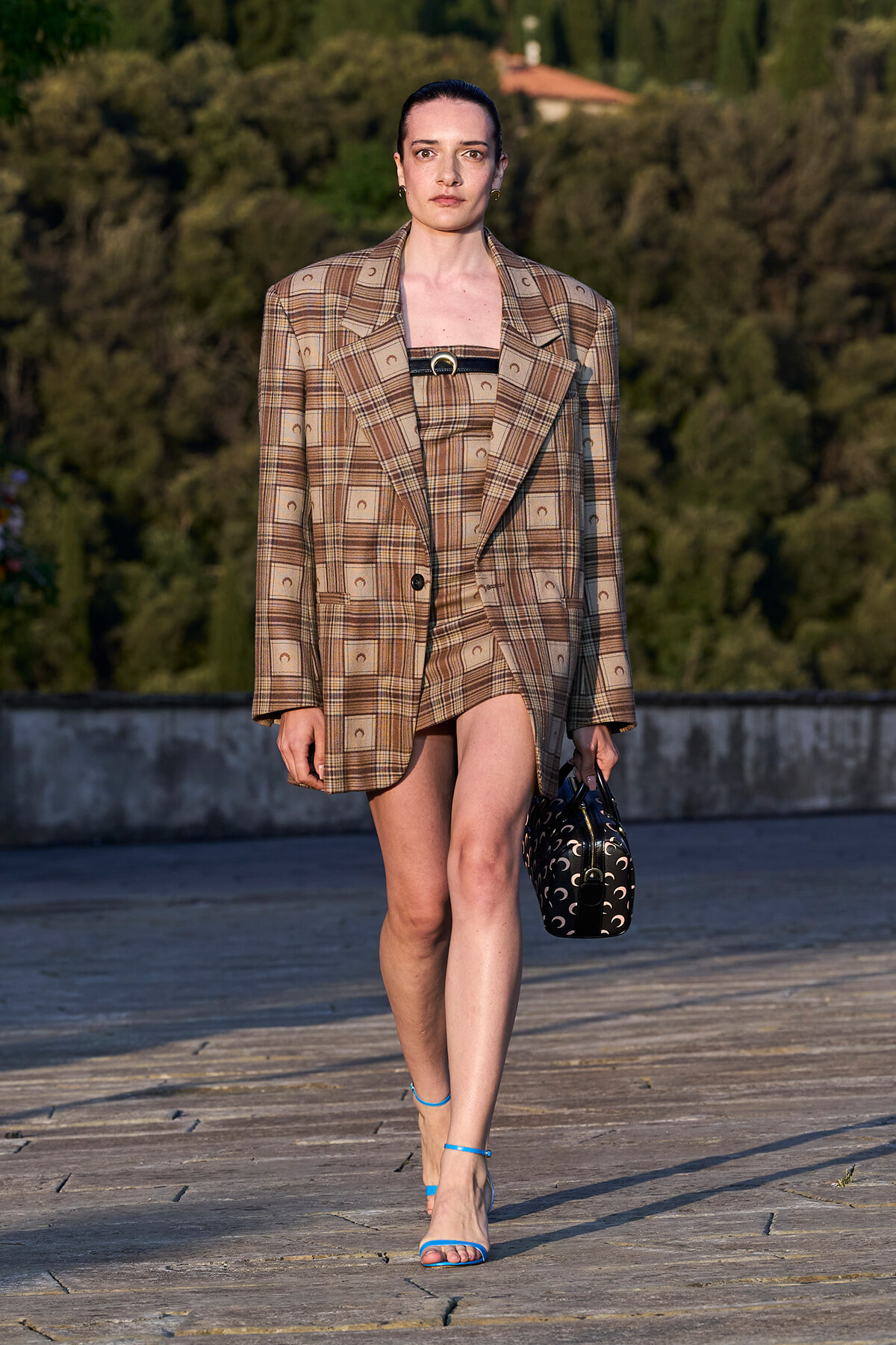 Model walking on a stone terrace in a brown plaid oversized blazer and matching skirt, blue strappy sandals, and a black patterned handbag.
