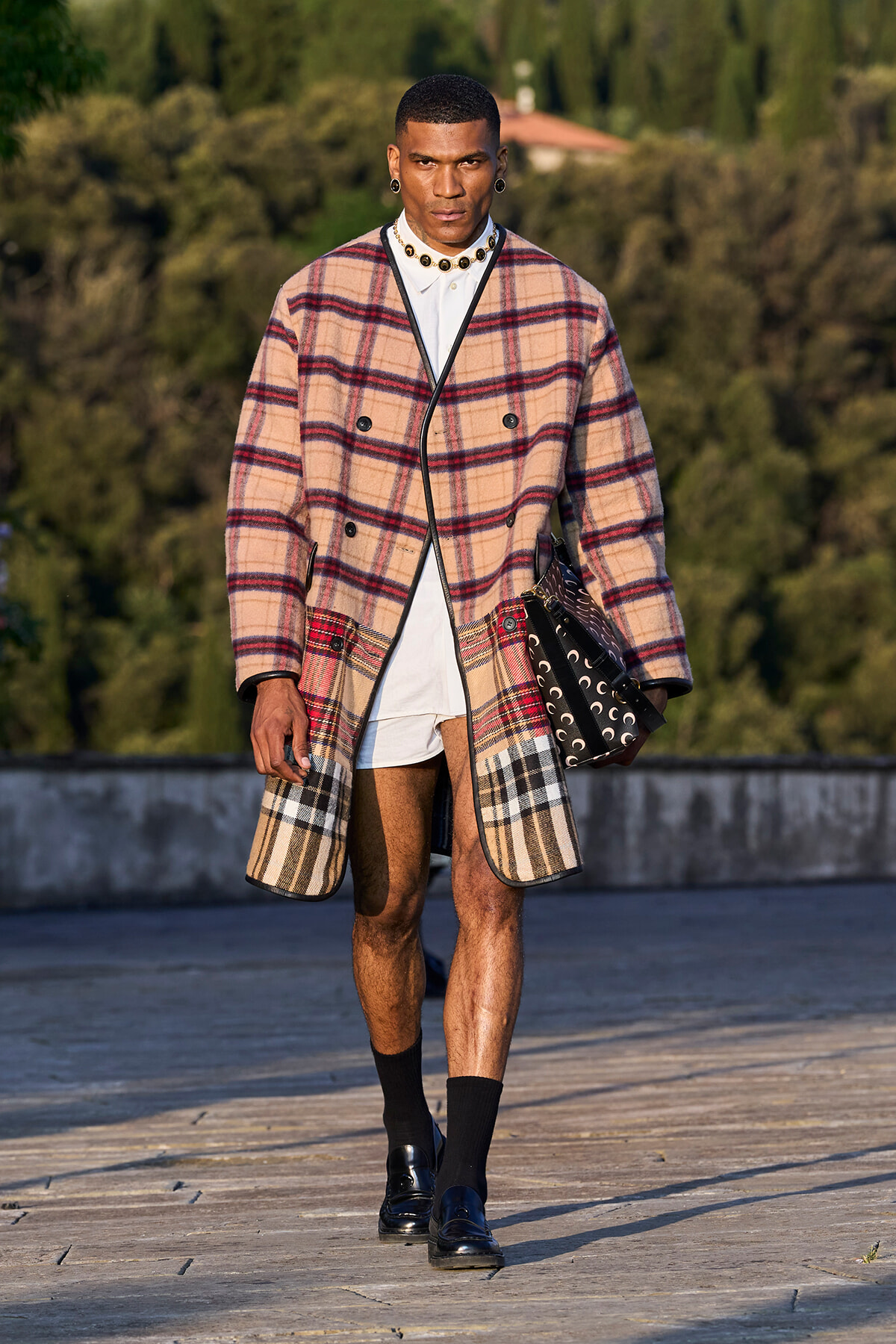 Male model on a runway wearing a beige plaid oversized coat with black trim, a white shirt and shorts, black socks and loafers, accessorized with earrings and a necklace, carrying a patterned bag.