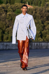 Male model walking outdoors in a pale blue striped shirt and shiny copper-brown patterned pants, carrying a light blue bag.