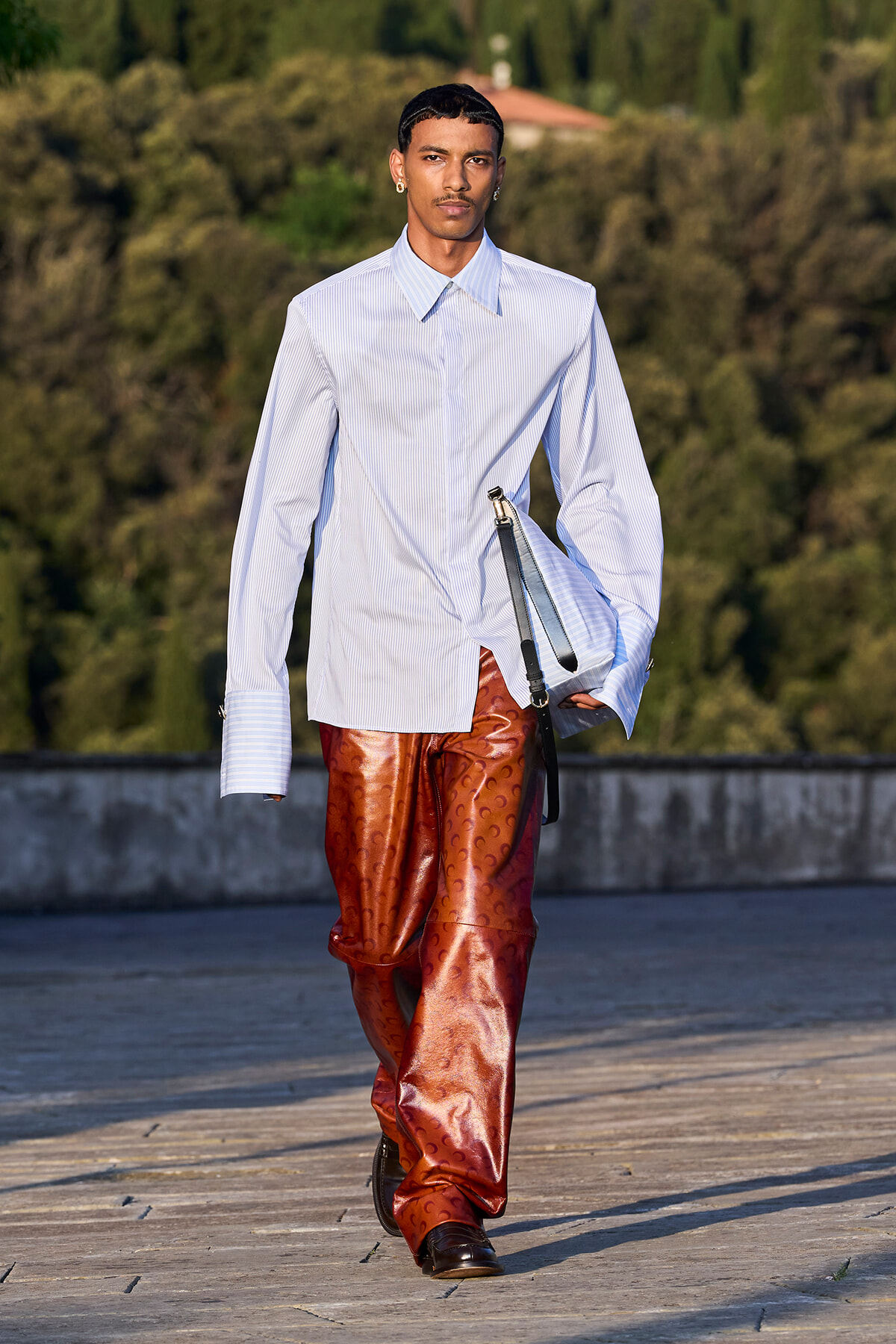 Male model walking outdoors in a pale blue striped shirt and shiny copper-brown patterned pants, carrying a light blue bag.