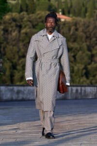 Man walking outdoors in a gray patterned trench coat with a belt, black shoes, and a brown briefcase in his left hand.