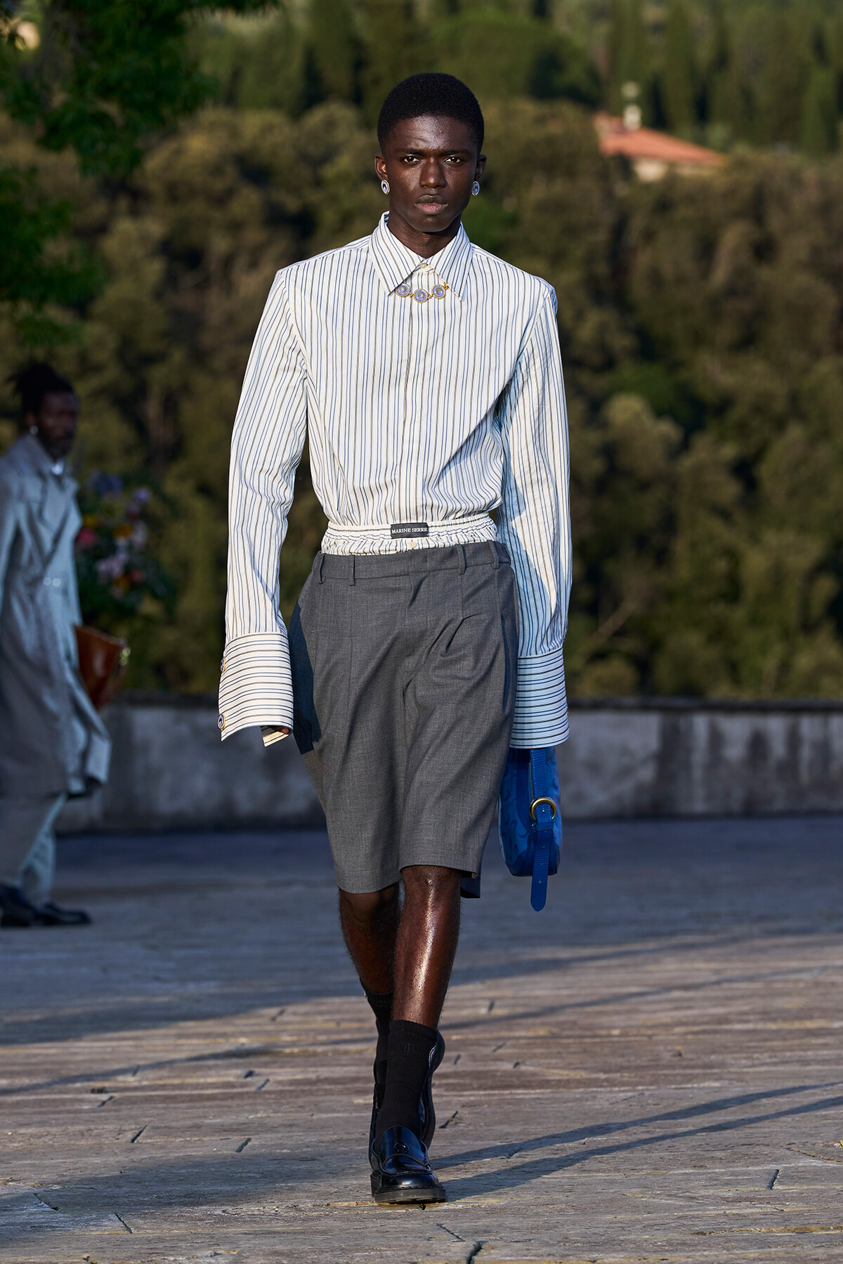 Male runway model in a striped shirt tucked into gray shorts, holding a blue handbag on a runway.