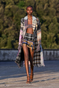 Model walking on an outdoor runway in a patchwork plaid coat over a brown bralette and matching plaid mini skirt, wearing black heeled sandals.