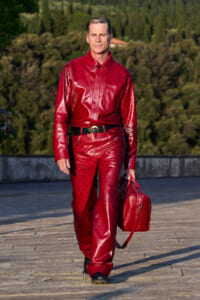 Man in a full red leather suit with belt walks across a stone terrace, carrying a matching red bag against a backdrop of trees and hills.