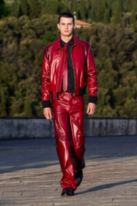 Male model in a shiny red leather suit and black shirt walks toward the camera on a rooftop terrace with greenery in the background.